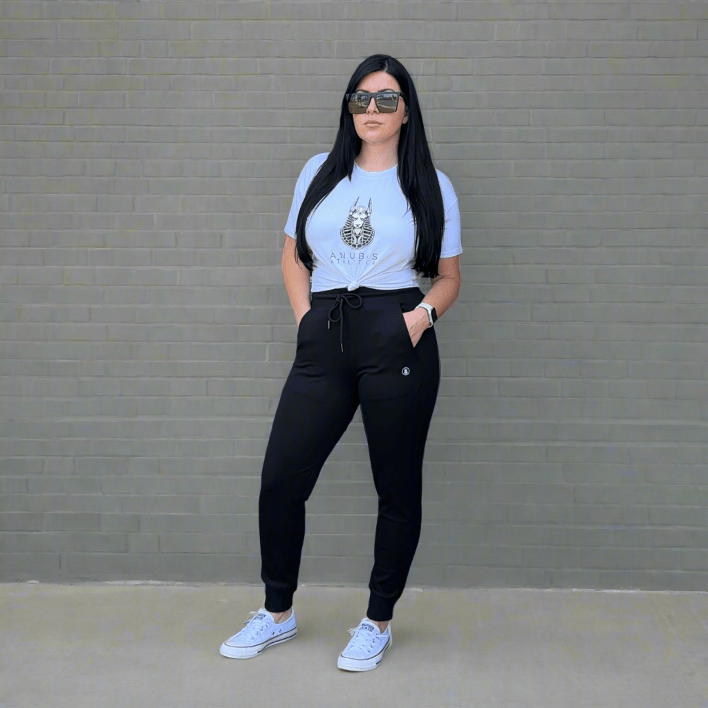 Woman wearing a white t-shirt with a graphic design, black pants, and white sneakers against a gray brick wall.