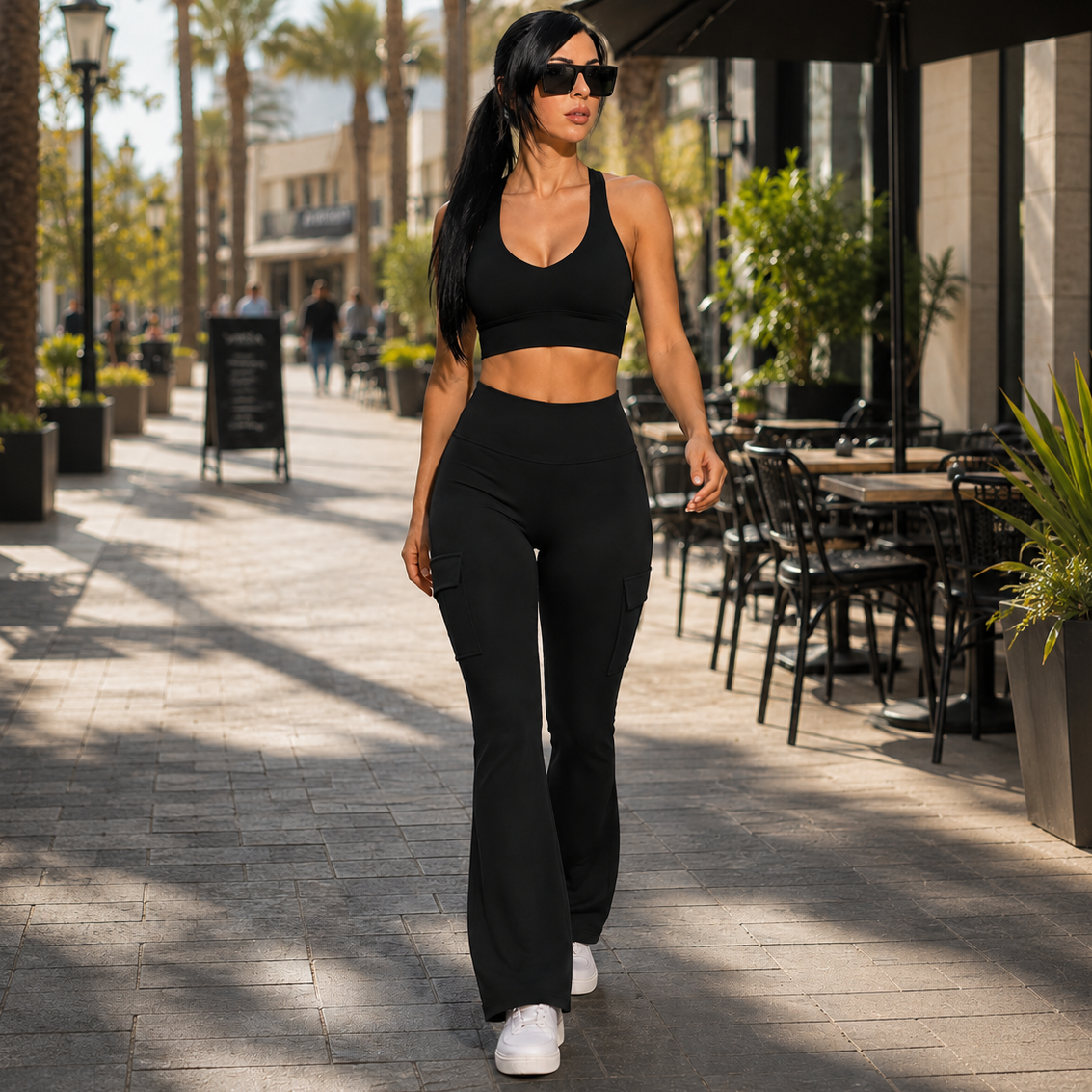 A woman walking outdoors on a sunny city sidewalk wearing a matching black activewear set consisting of a halter crop top and high-waisted flared leggings, paired with white sneakers.