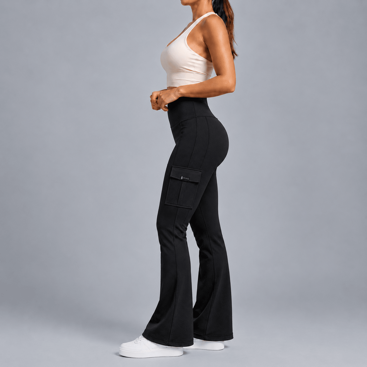 A woman in a side profile wearing high-waisted black flared cargo pants, a light beige tank top, and white sneakers against a solid gray studio background.