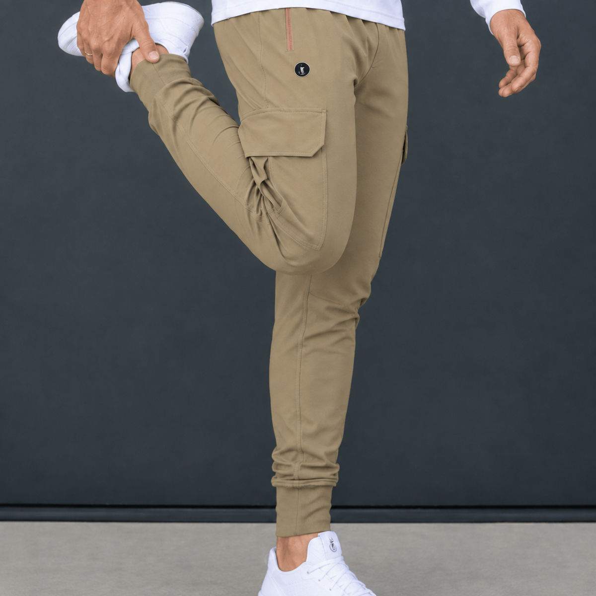 A person wearing khaki cargo jogger pants, shown from the waist down, stretching their leg while wearing white sneakers against a dark gray studio background.