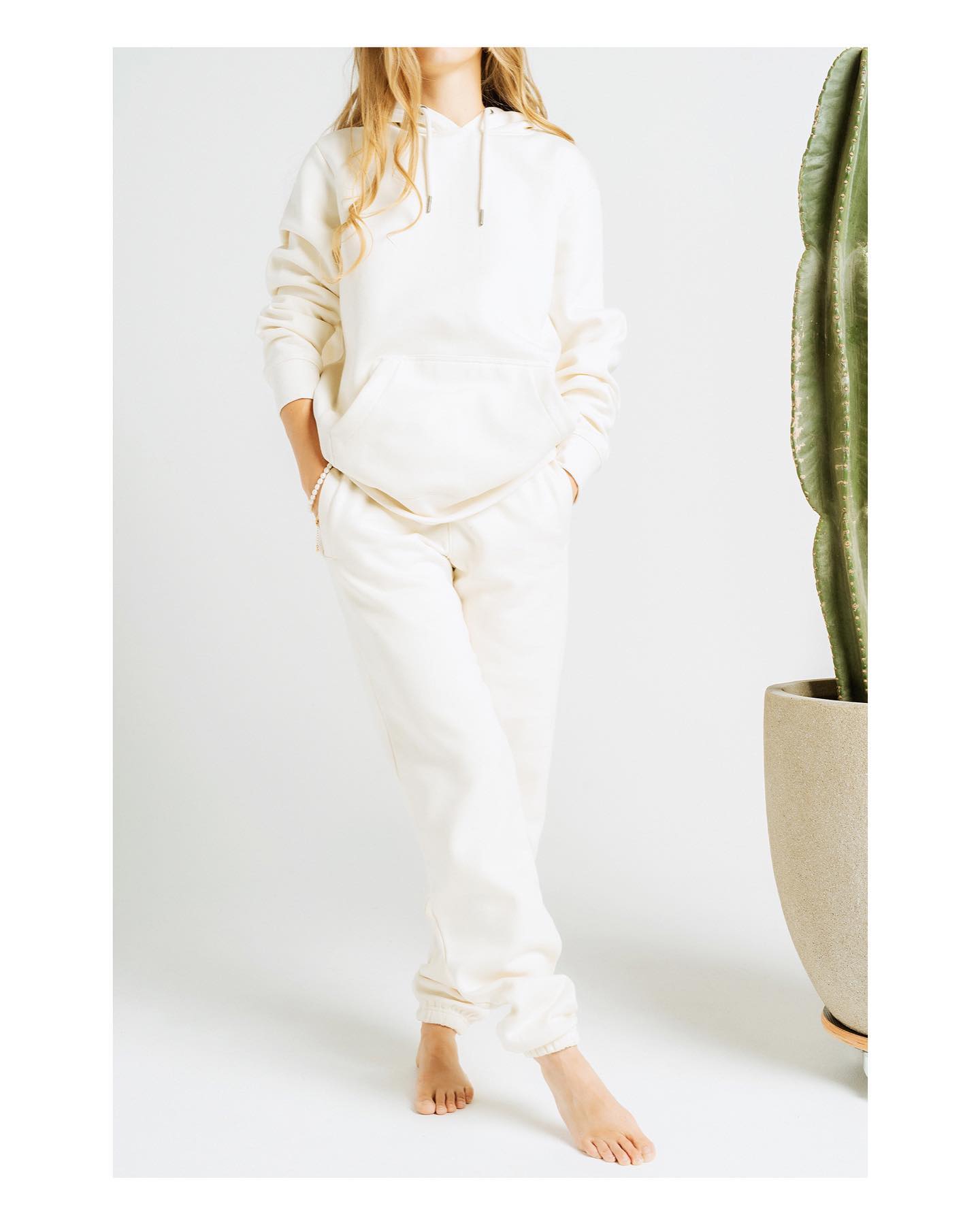 Blonde Caucasian female model standing next to a cactus wearing a cream colored pull over hoodie and a pair of matching sweatpants.