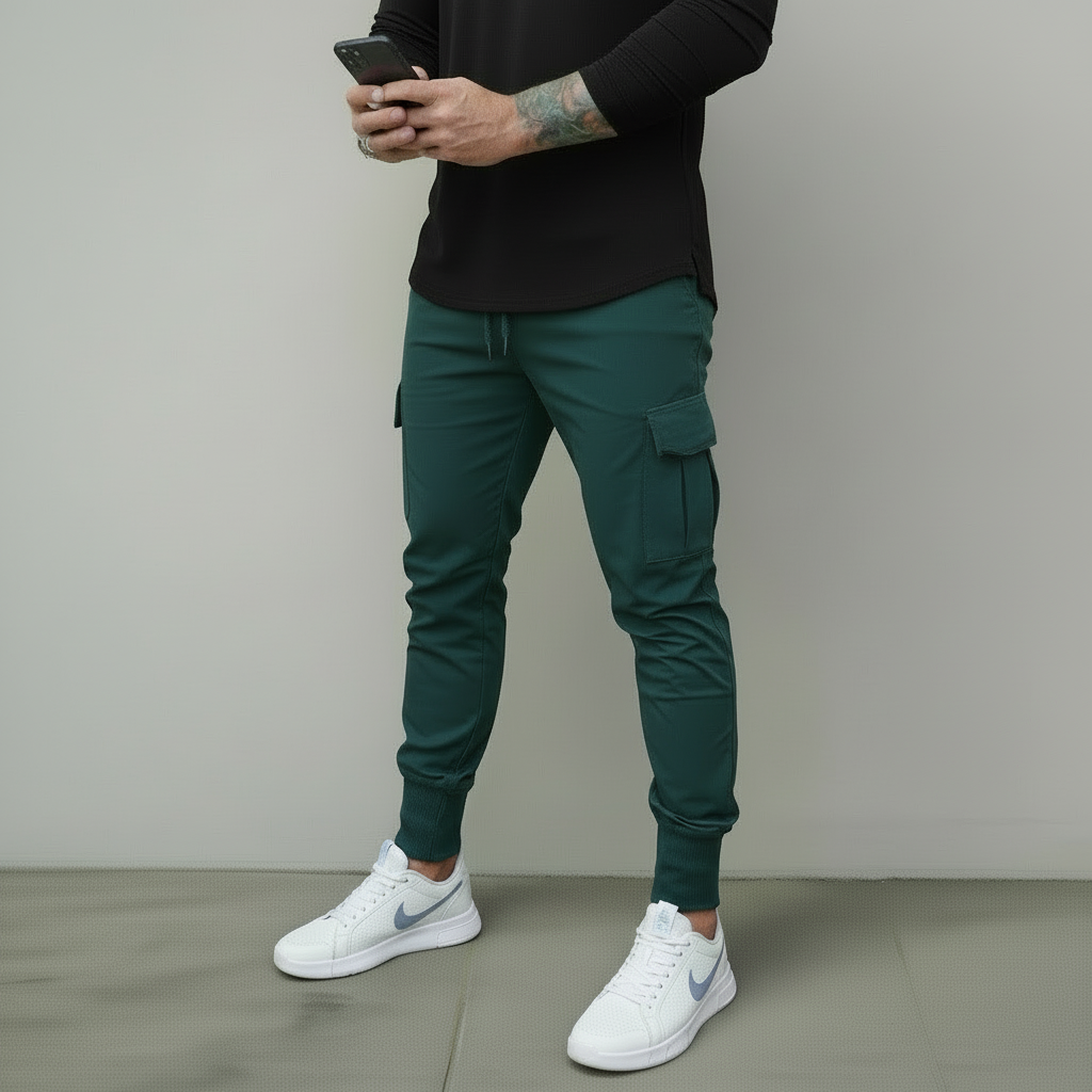 Person wearing a black long-sleeve shirt, teal cargo pants, and black sneakers against a beige wall.