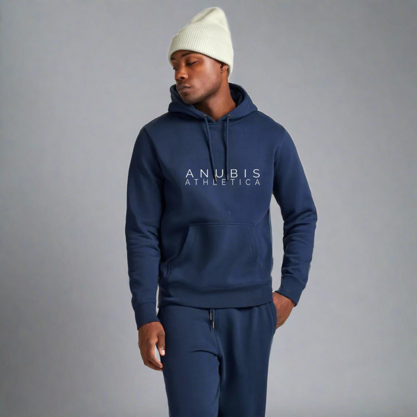 African American male model standing with a white beanie on his head wearing a blue pull over hoodie with the name Anubis Athletica printed in the center. Also wearing a pair of matching sweatpants