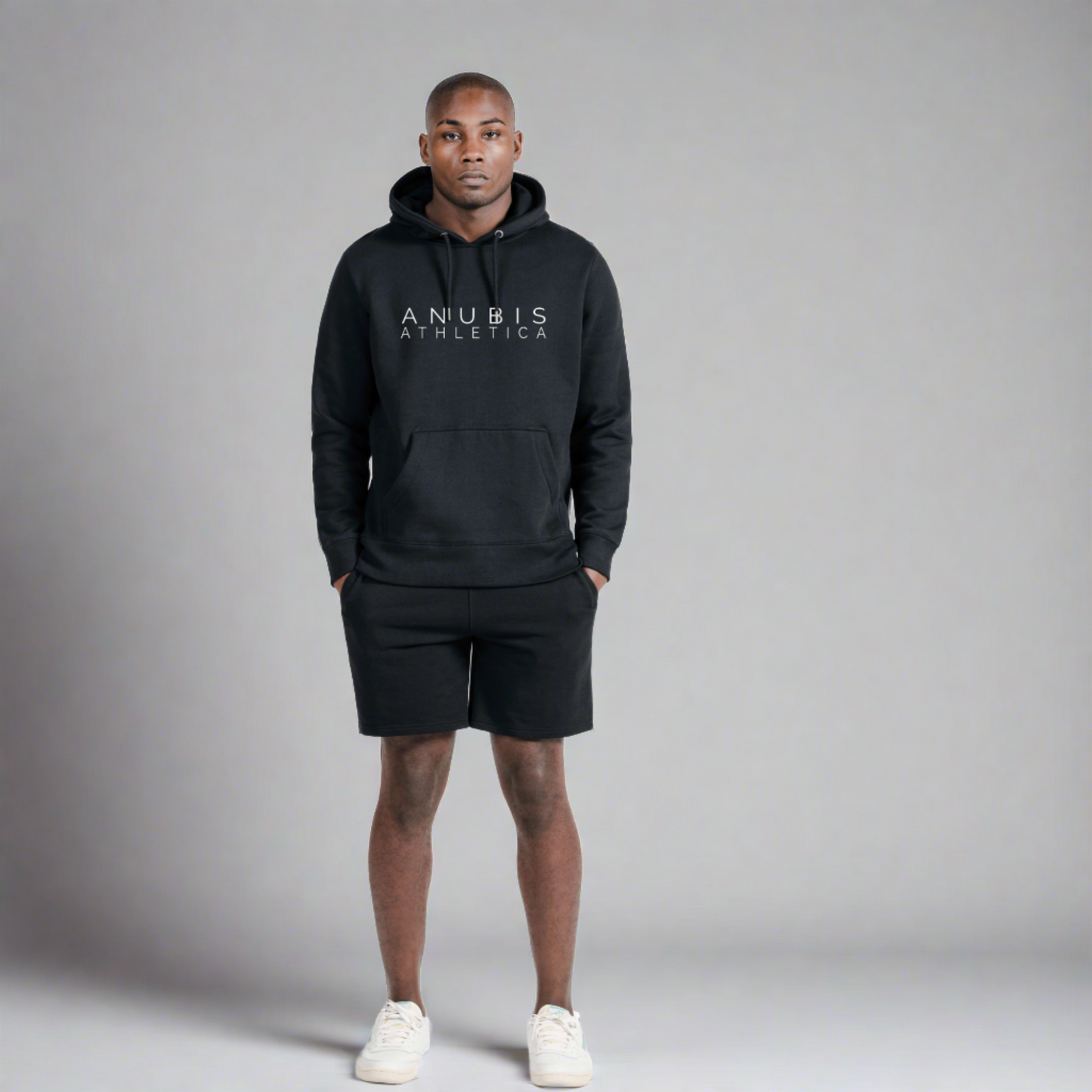 Black male mode wearing a pull over hoodie with the name Anubis Athletica printed in the center and also wearing a pair of matching sweat shorts