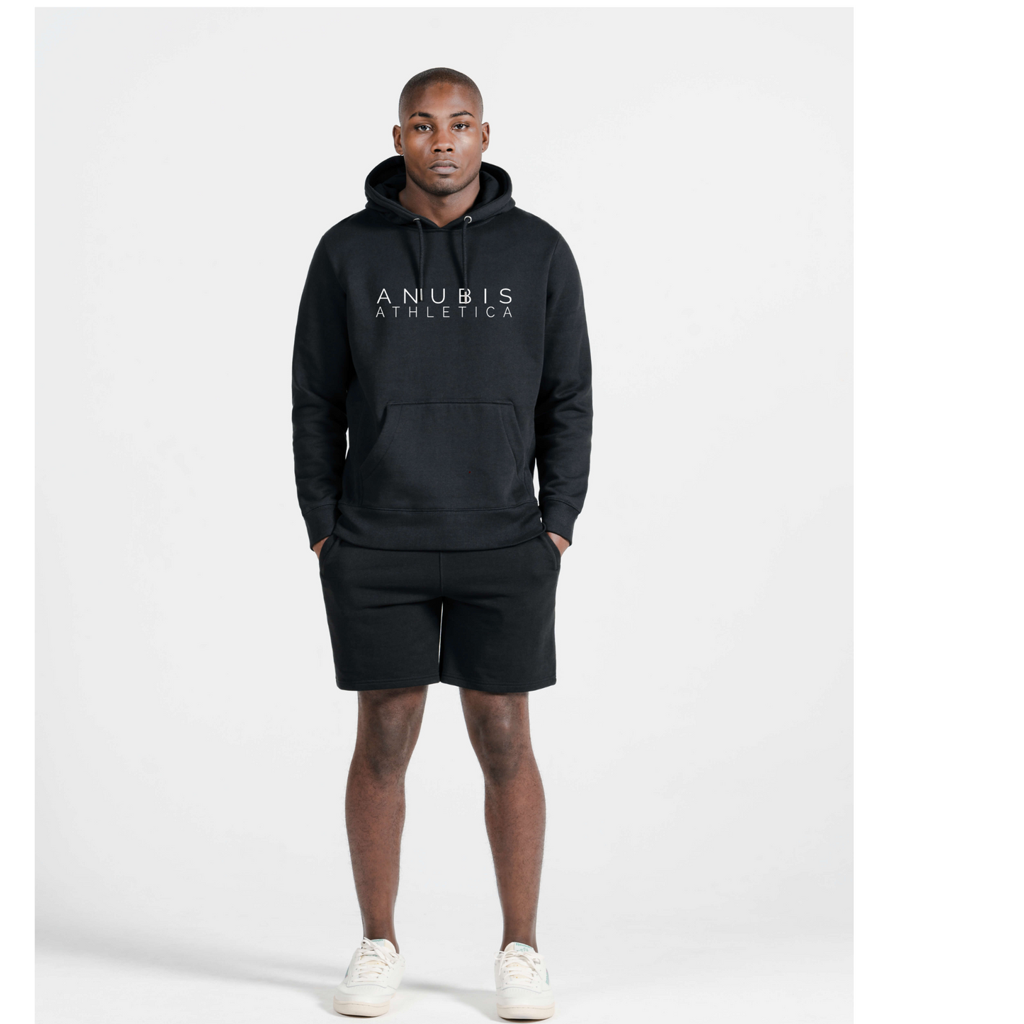 Black male mode wearing a pull over hoodie with the name Anubis Athletica printed in the center and also wearing a pair of matching sweat shorts