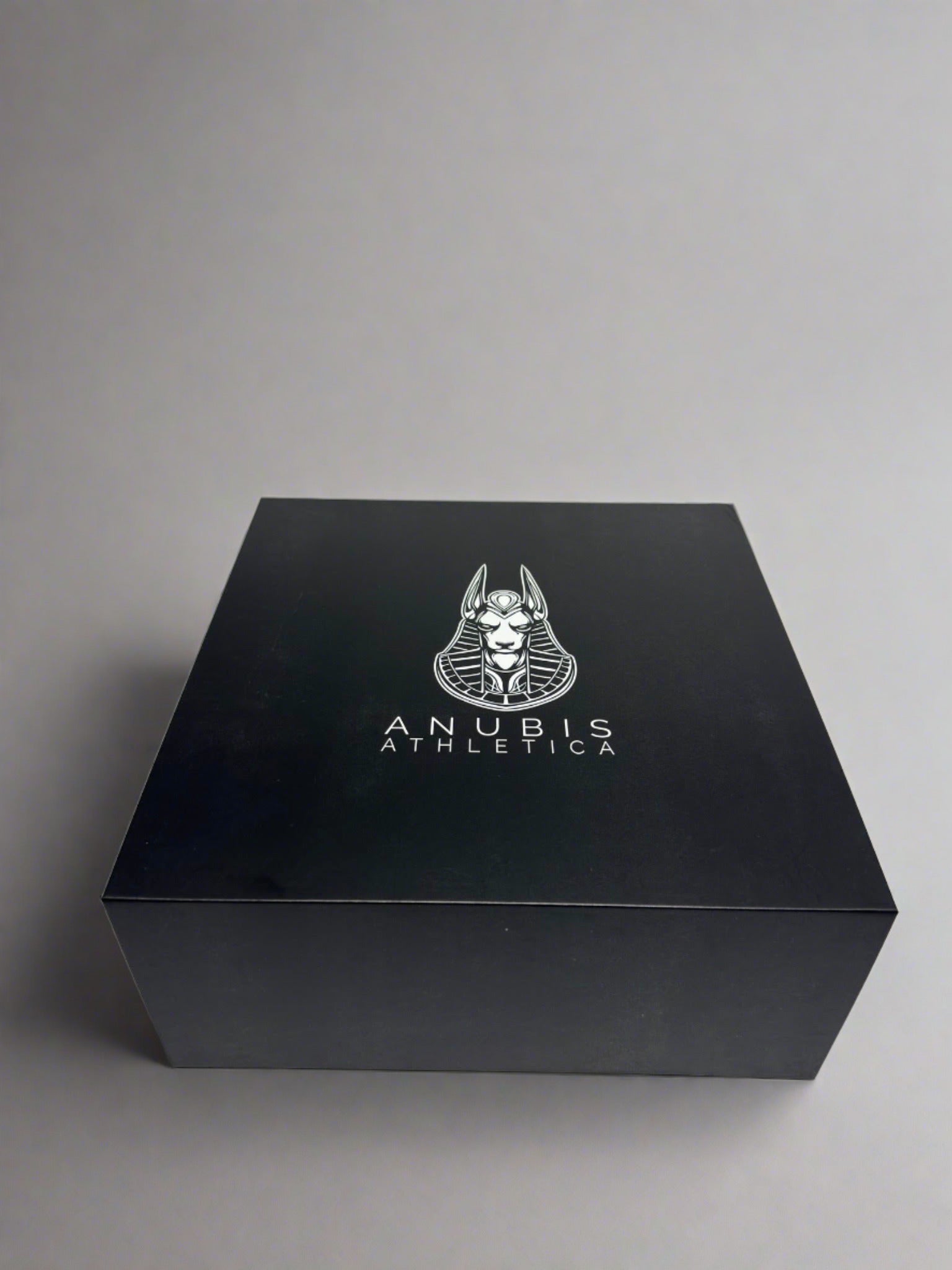 An example of the box the apparel is shipped in. The box is shiny black with a print of the Anubis Athletica logo printed in the center which includes the picture of Anubis and the name of the company below it.