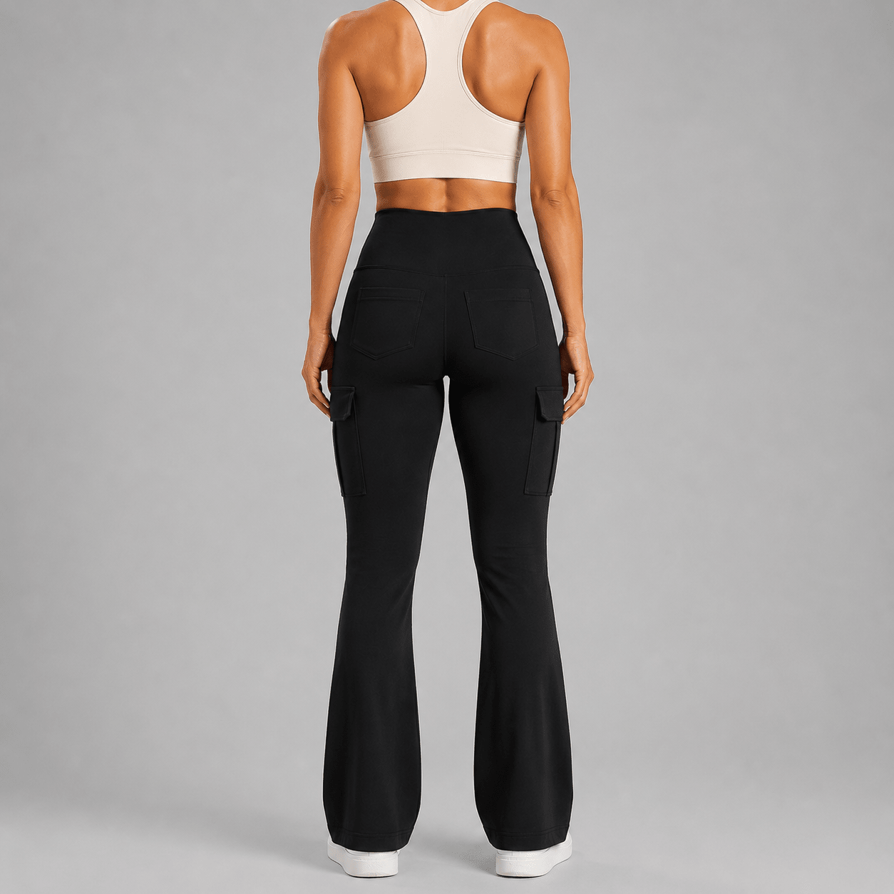 Rear view of a model wearing black high-waisted flared cargo pants and a cream sports bra against a solid gray studio background.