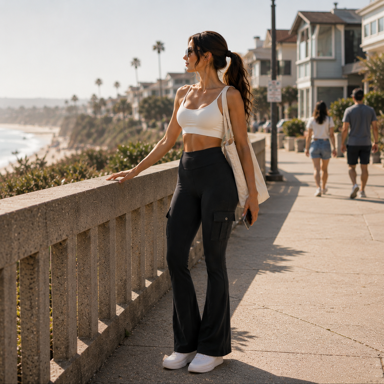A woman stands on a sunny beach boardwalk wearing a white ribbed tank top and black cargo flare pants, with a white shoulder bag and white sneakers.