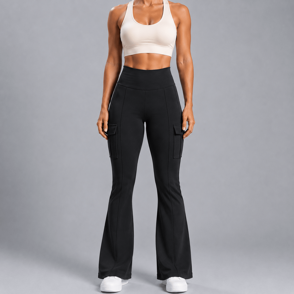 A model wearing black high-waisted flared cargo pants with side pockets, paired with a beige sports bra and white sneakers, standing against a solid gray studio background.