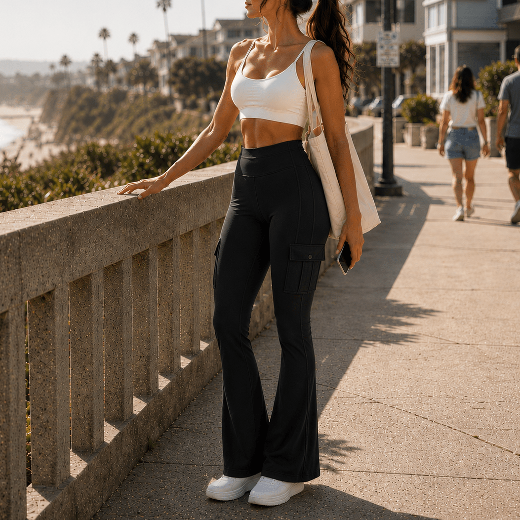 A woman stands on a sunny beach boardwalk wearing a white sports bra and black flared cargo pants, with a white tote bag and sunglasses. The background shows a coastal walkway, ocean, palm trees, and houses.
