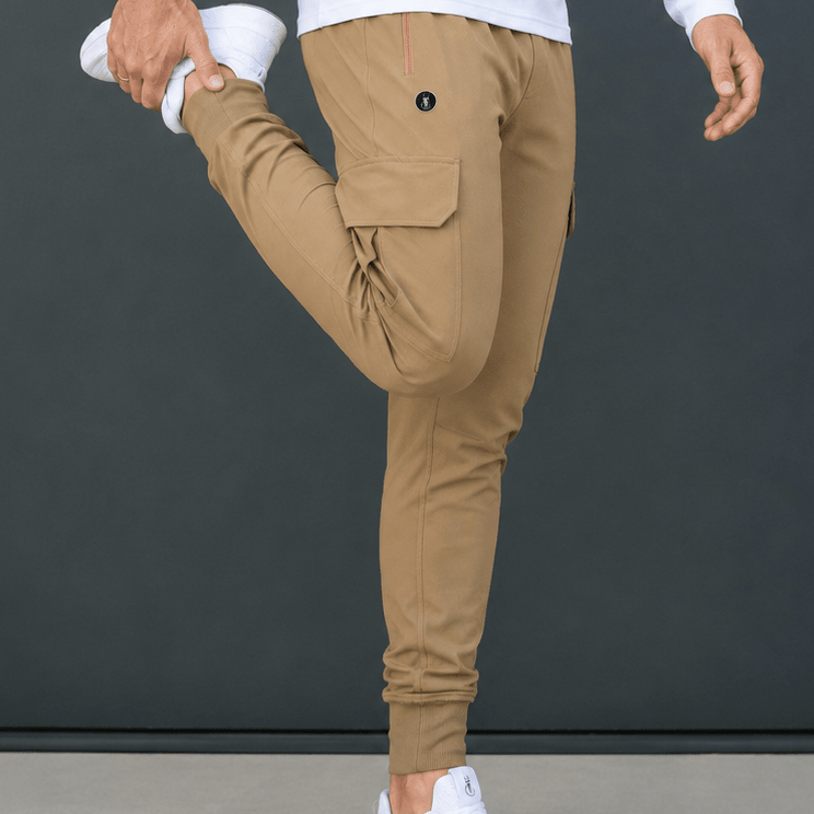 A person wearing tan cargo jogger pants, a white long-sleeve shirt, and white sneakers is shown from the waist down, performing a leg stretch against a dark gray studio background.