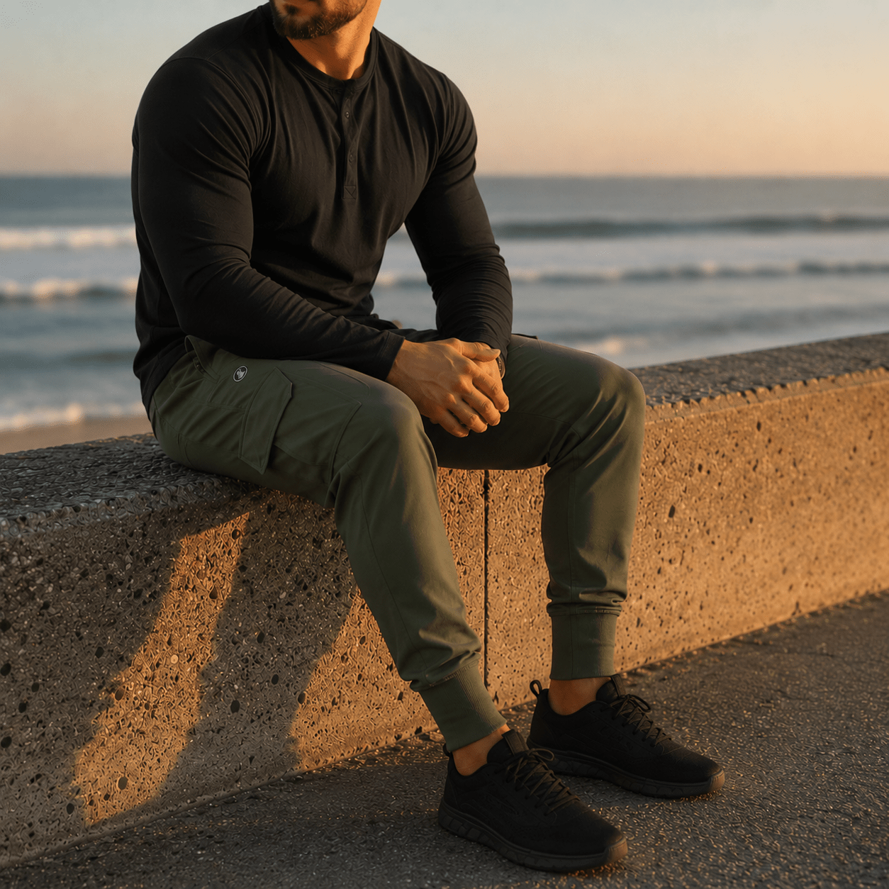 A man sitting on a concrete seawall by the ocean at sunset, wearing a black long-sleeve henley shirt, olive green cargo jogger pants, and black sneakers.