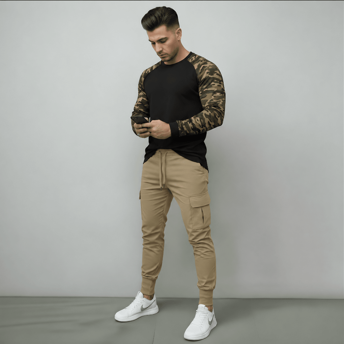 A man modeling a long-sleeve raglan shirt with a black body and camouflage sleeves, paired with beige cargo jogger pants and white sneakers, standing against a plain gray studio background.