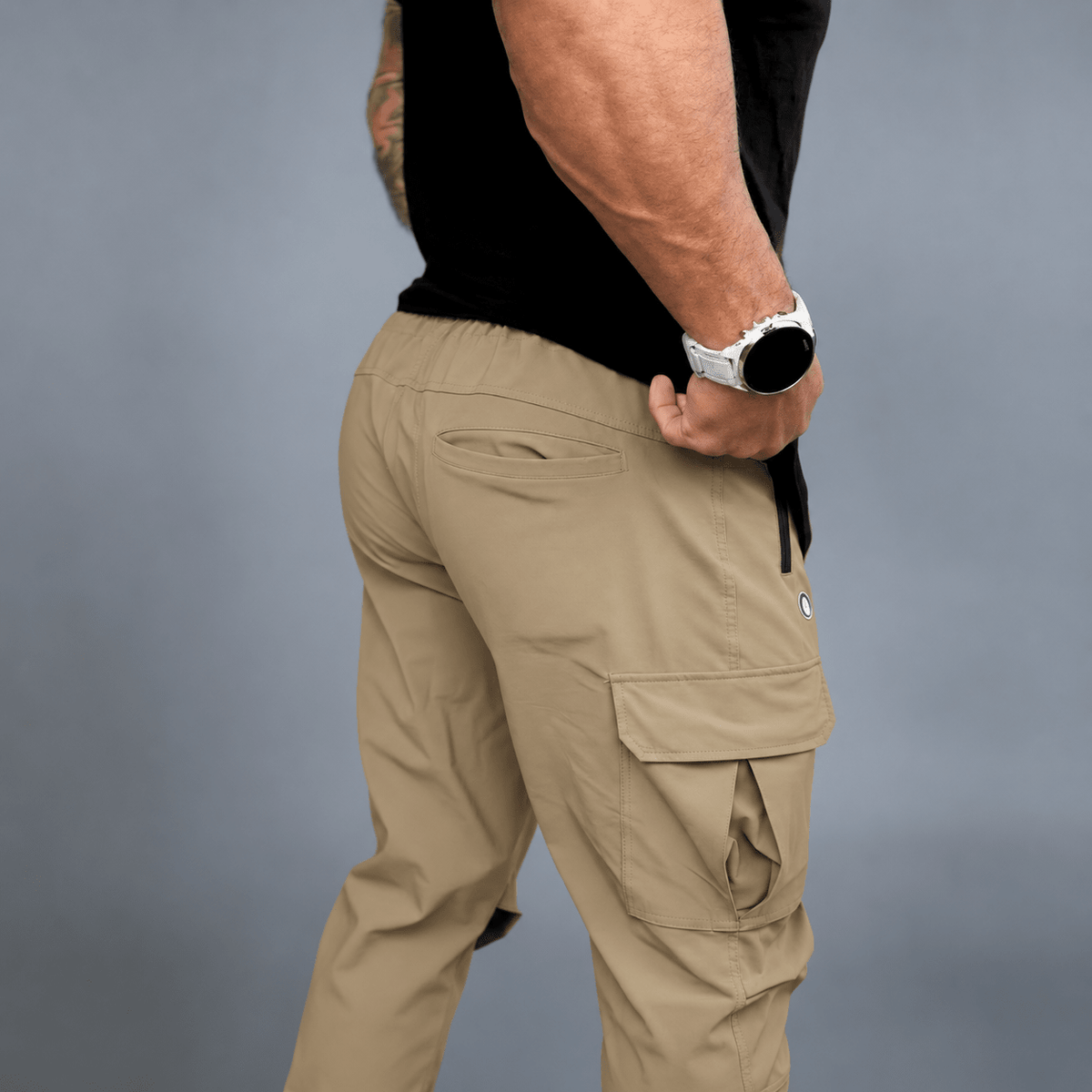A man wearing khaki cargo pants with a black t-shirt and a white smartwatch, shown from a side-back angle against a gray studio background.