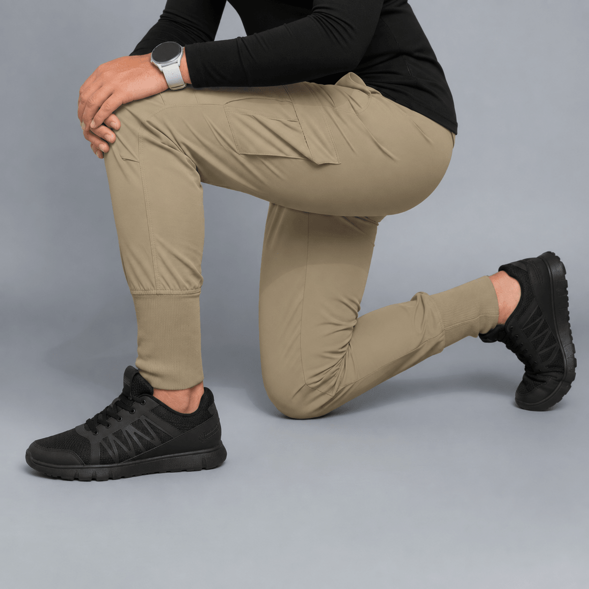 A model kneels in a studio setting wearing khaki cargo jogger pants, a black long-sleeve top, and black sneakers against a solid gray background.