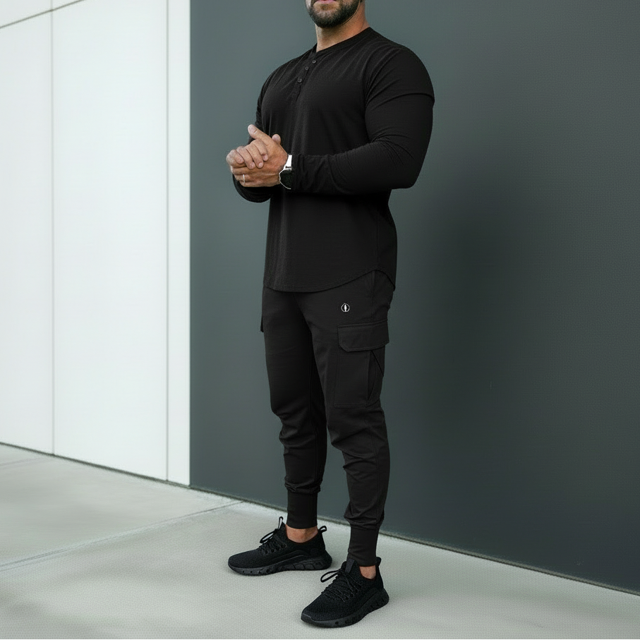 Man wearing a black outfit standing against a gray wall.