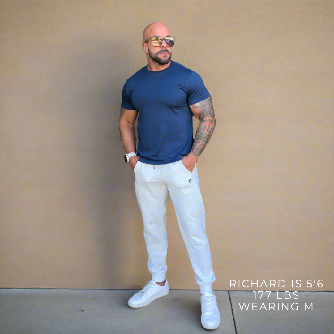 Man wearing a blue t-shirt, white pants, and white sneakers against a beige wall.