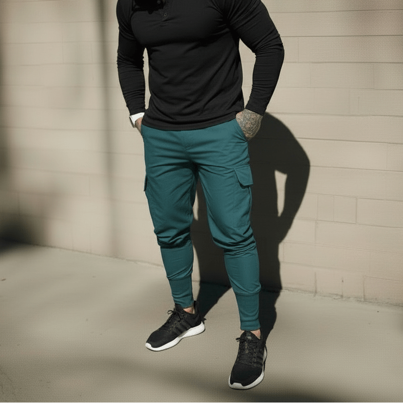 Man wearing a black shirt, teal cargo pants, and black sneakers against a concrete wall.