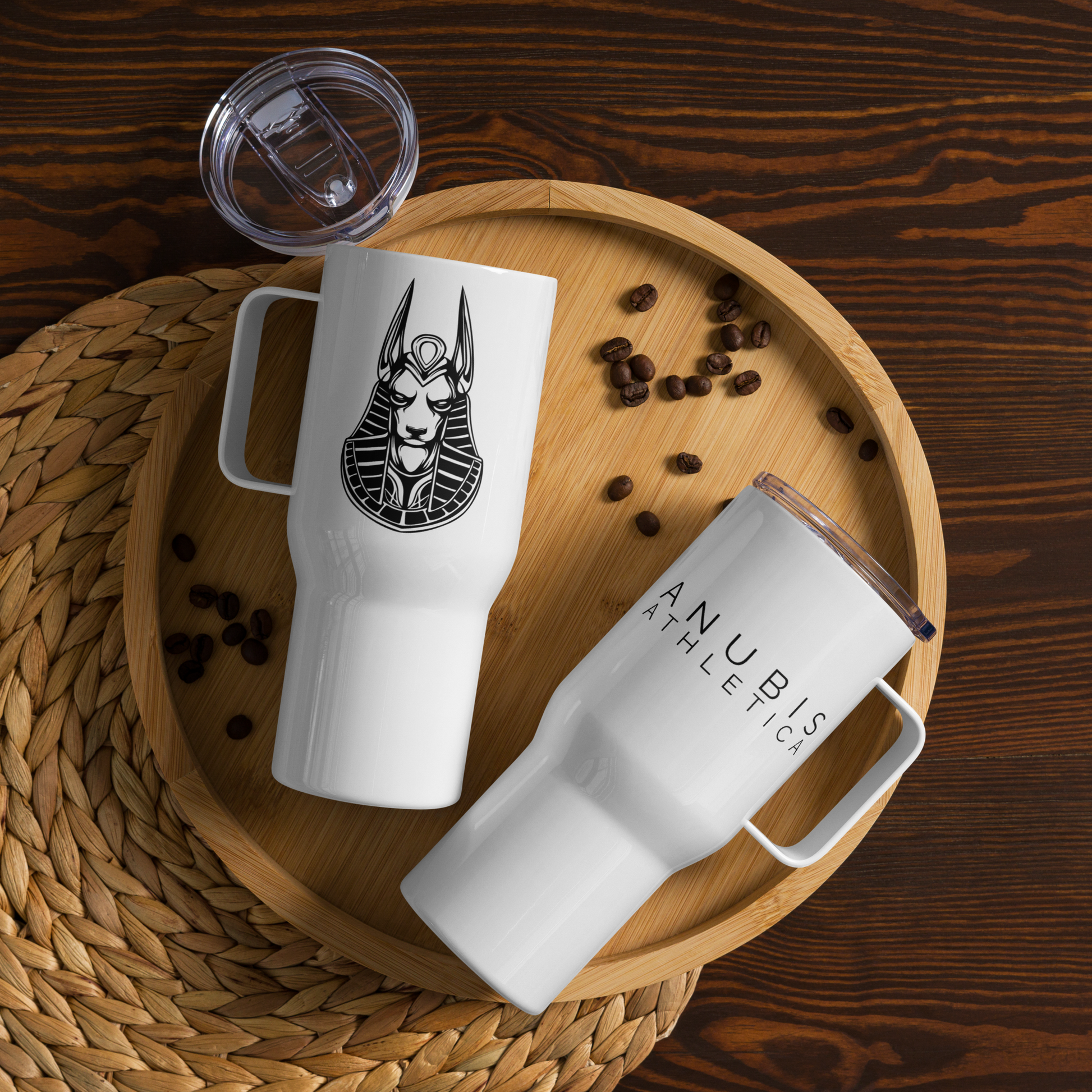 two white tumblers with handles. One shows the name Anubis Athletica on the front and one shows a picture of Anubis on the side. They are on a wooden platter with a few coffee beans around them